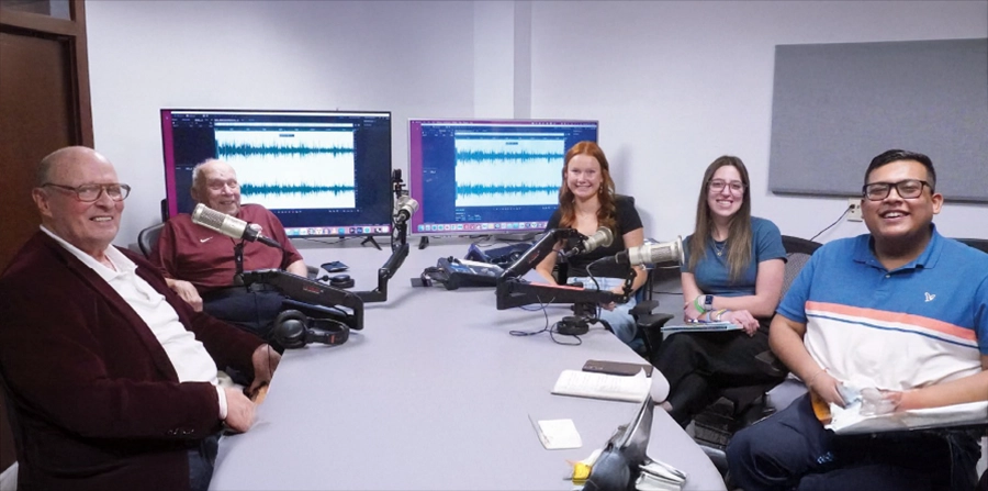 Five people seated around a table with microphones in a recording studio, two screens showing audio waveforms in the background.