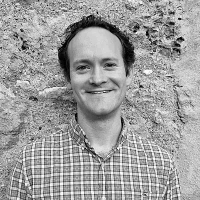 Man smiling in front of a textured stone wall, wearing a plaid shirt. Black and white image.