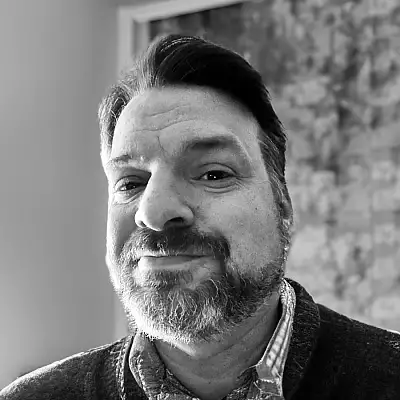 A black-and-white portrait of a man with short hair and a beard, wearing a collared shirt and sweater, looking at the camera.