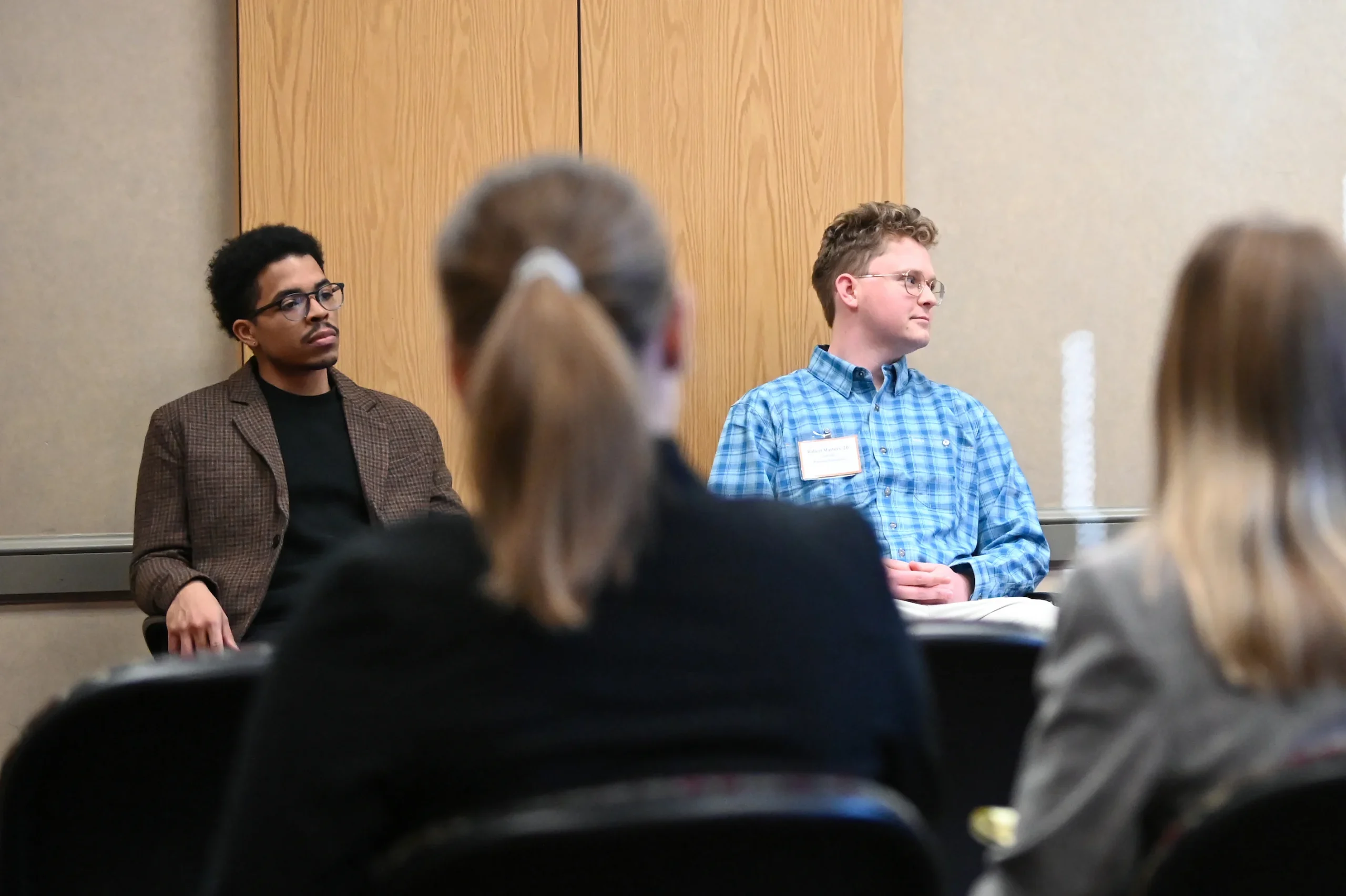 A group of people sit in chairs during a meeting. Three men are at the front, and two women in the foreground are seen from behind, with one using a laptop.