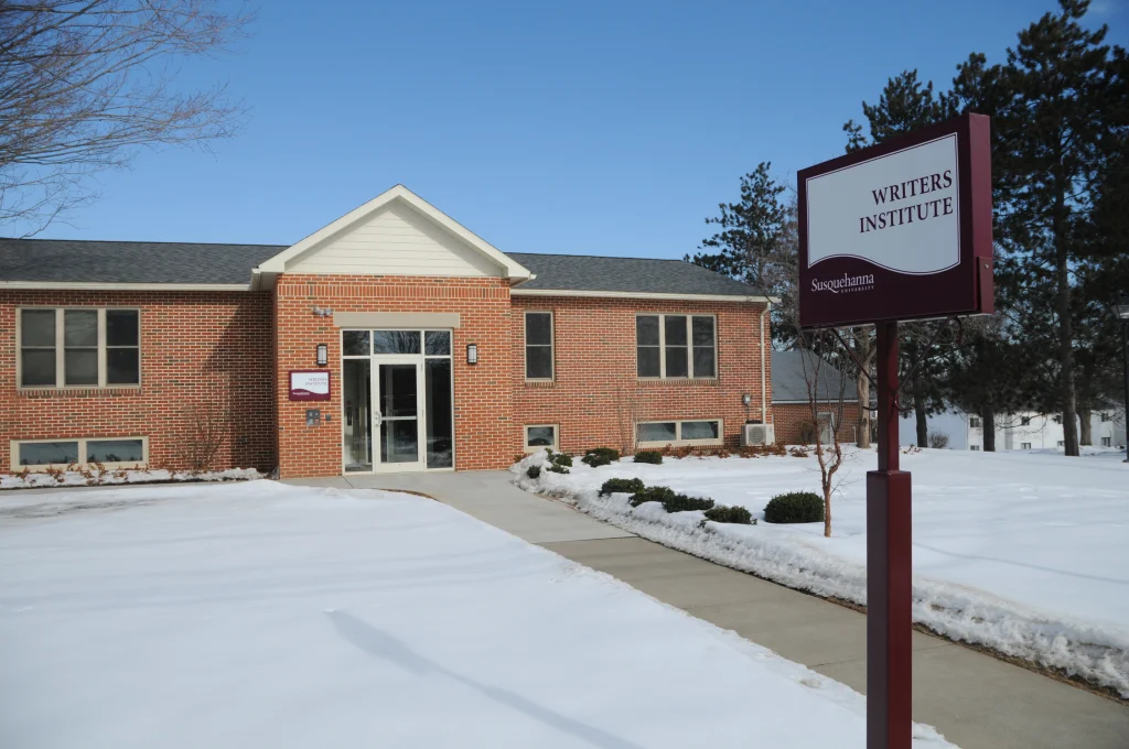 Red-brick building labeled "Writers Institute" with a sign and snow-covered ground. Clear blue sky in the background.