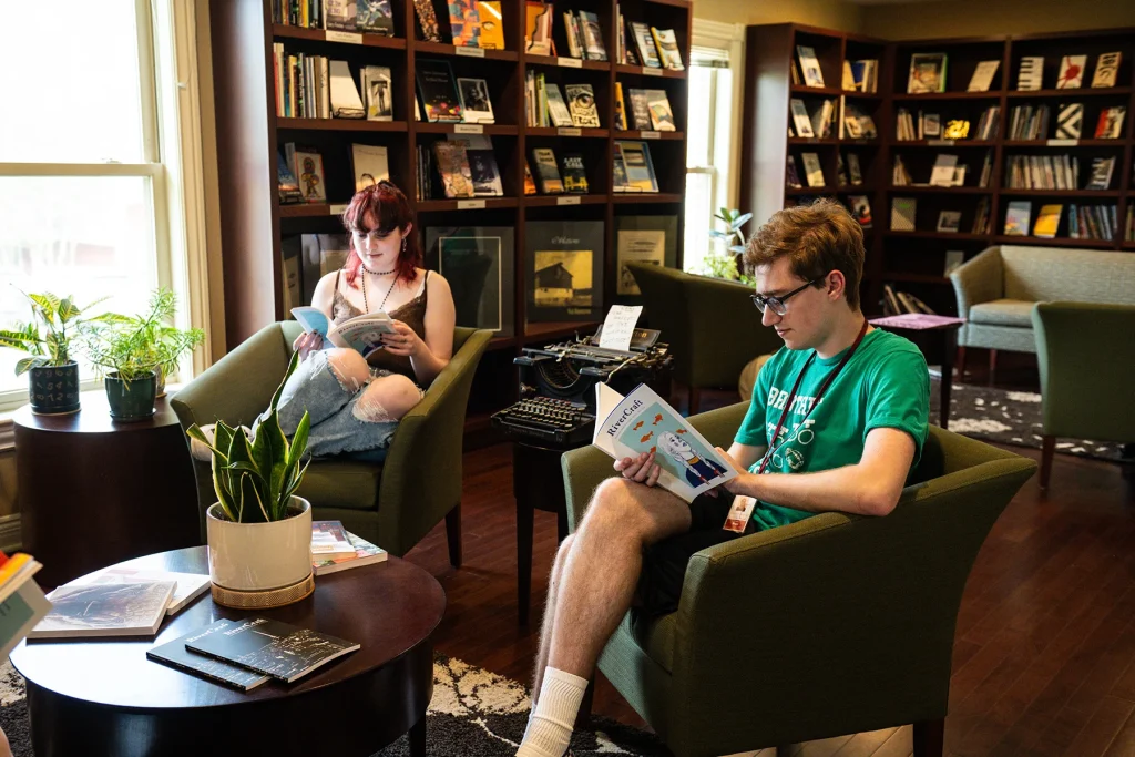 Two people sitting and reading in a cozy library with wall shelves filled with books and records. A typewriter and a potted plant are on tables nearby.