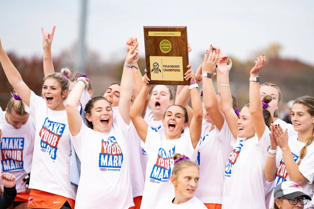 A group of jubilant young athletes wearing "Make Your Mark" shirts celebrate winning a field hockey championship, holding up a trophy.