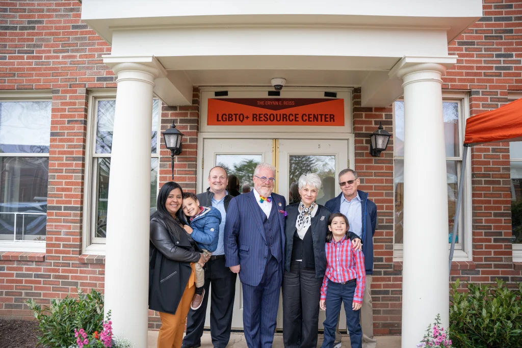 A group of seven people, including adults and children, stands in front of a building with a sign reading "LGBTQ+ Resource Center." The background has brick walls and a white doorway.