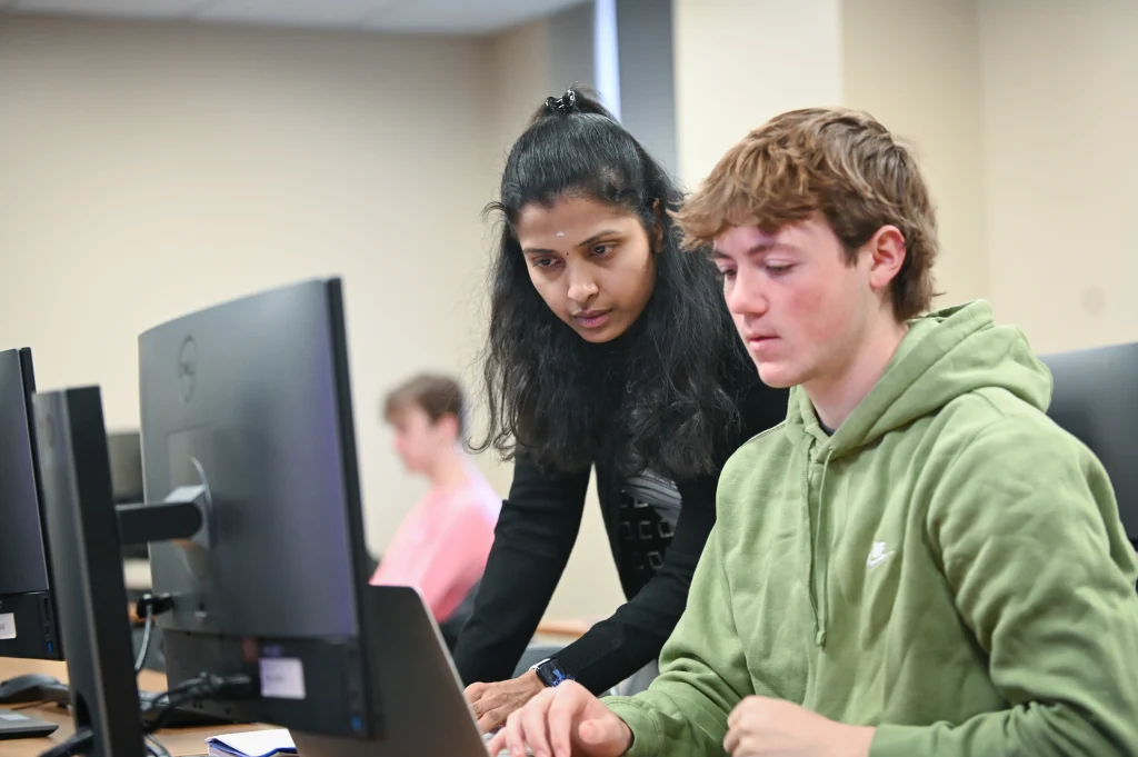 A woman assists a young man working on a computer in a classroom setting. Other students are visible in the background, focusing on their screens.