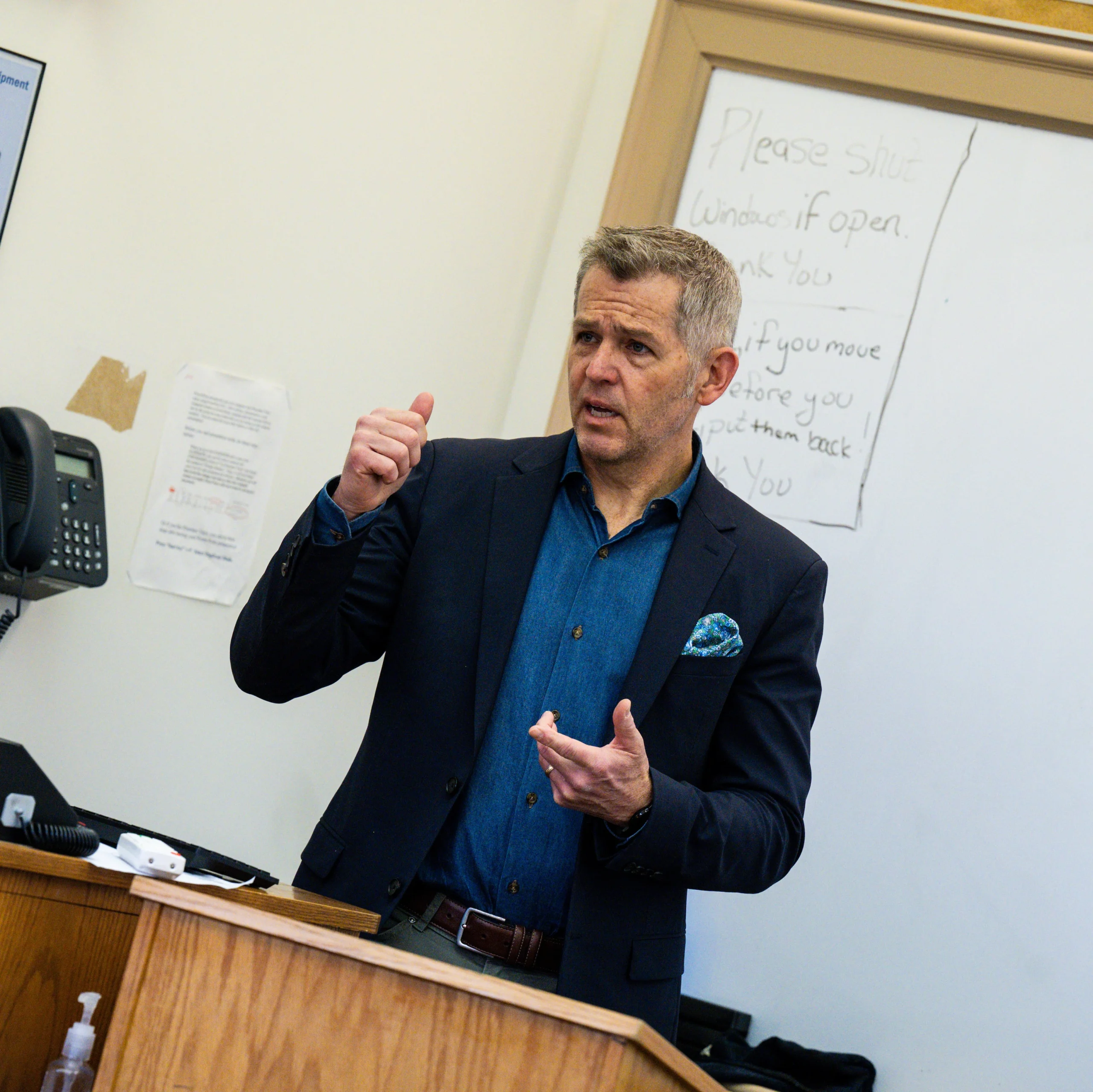 A man in a blazer gestures while speaking at a podium in a classroom. A whiteboard with handwritten notes is in the background.
