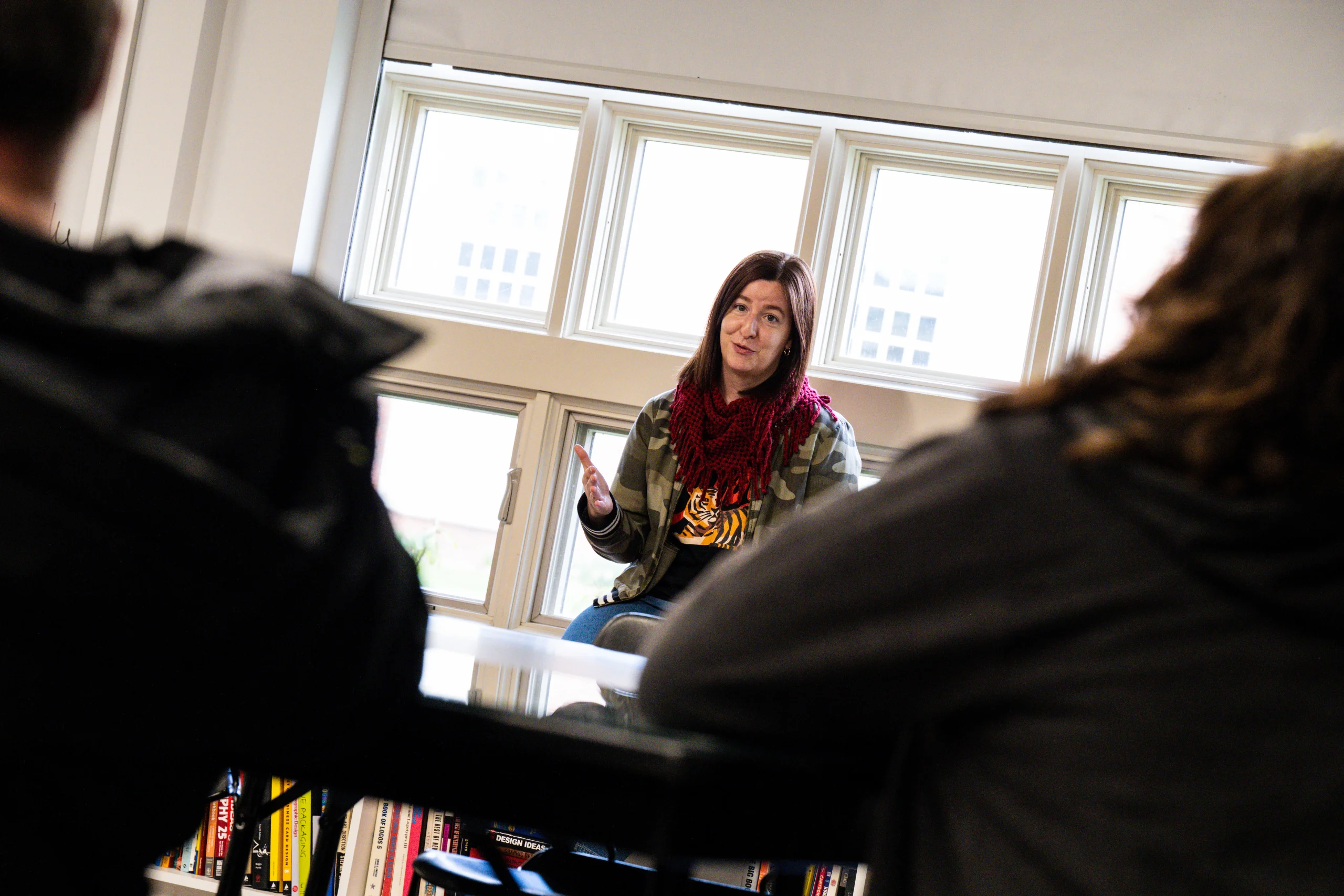 A person sits on a table in front of a window, talking to a small group. Shelves filled with books are visible below the table.