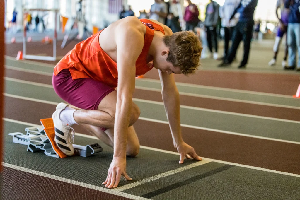 An athlete in an orange tank and maroon shorts kneels at the starting line on an indoor track, preparing to sprint.