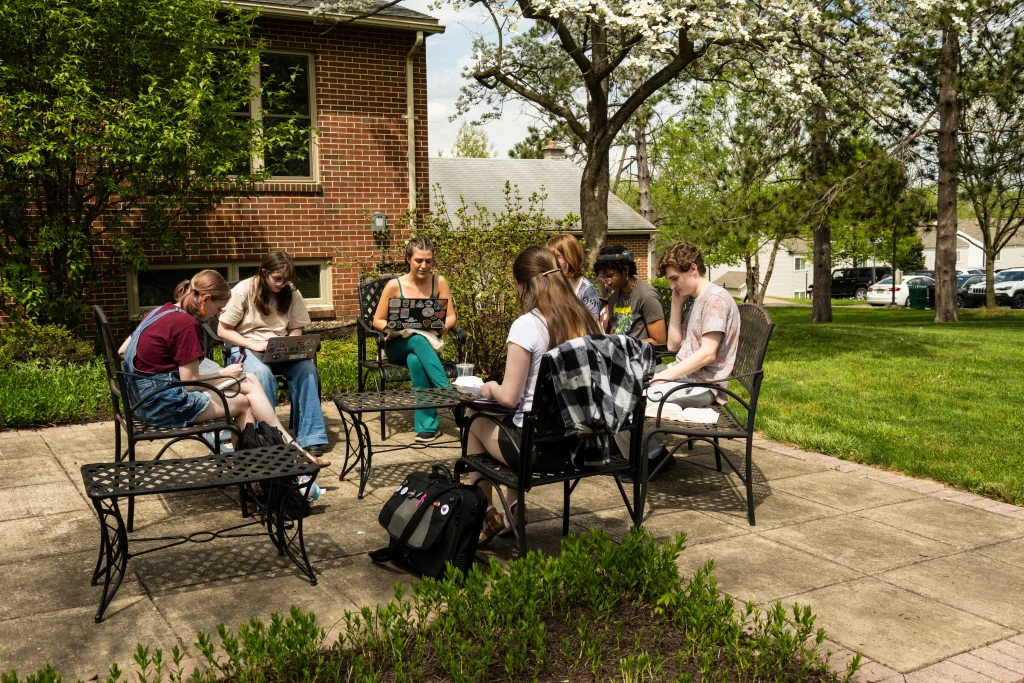 Seven people sit outdoors around a table, engaging in conversation on a patio surrounded by greenery and brick buildings.