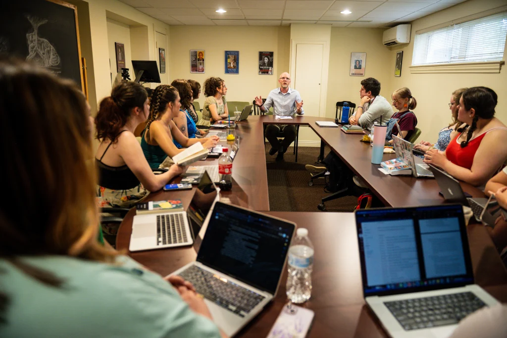 A group of people sit around a conference table with laptops and notebooks while one person speaks. A chalkboard and various items are visible in the room.