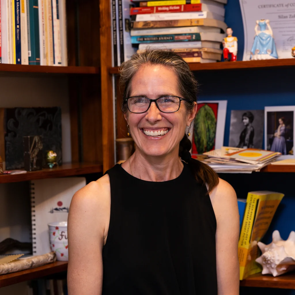 Smiling woman with glasses and a black sleeveless top sits in front of a wooden bookshelf filled with books and decorations.