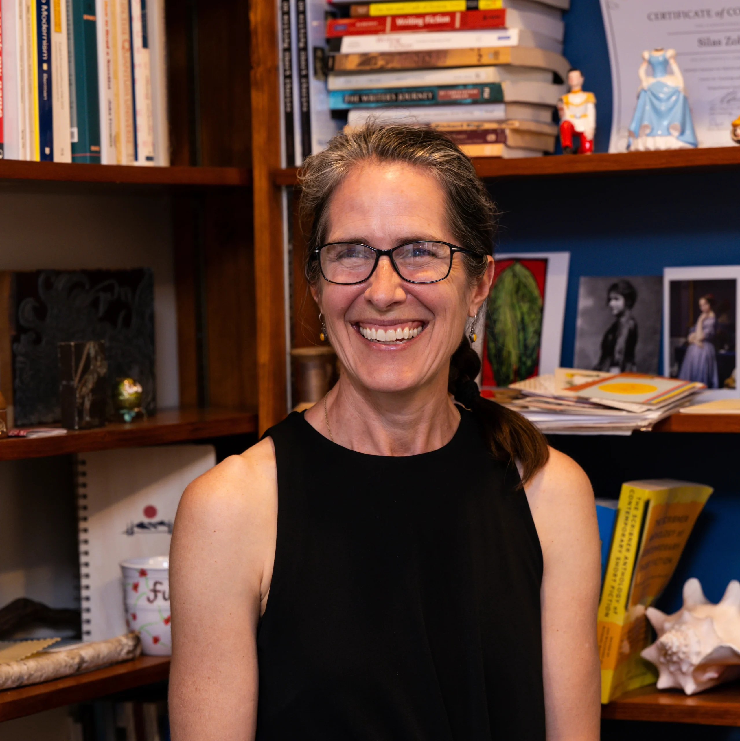 Smiling woman with glasses and a black sleeveless top sits in front of a wooden bookshelf filled with books and decorations.